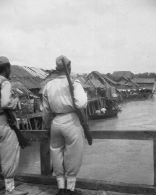 KNIL militairen staan op wacht op een brug en kijken uit over vissersdorp. Fotograaf: Van Buren / DLC