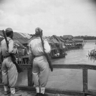 KNIL militairen staan op wacht op een brug en kijken uit over vissersdorp. Fotograaf: Van Buren / DLC